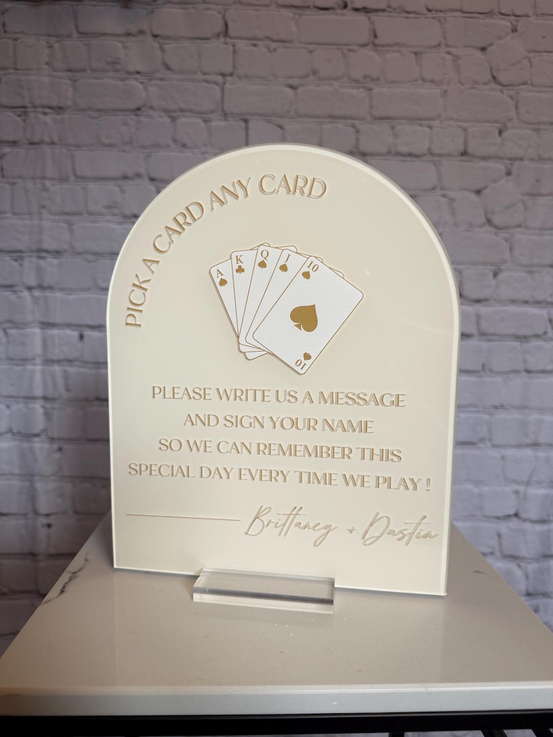 May include: An arched cream-colored acrylic sign featuring gold text and a graphic of five playing cards. The text reads: PICK A CARD ANY CARD, PLEASE WRITE US A MESSAGE AND SIGN YOUR NAME SO WE CAN REMEMBER THIS SPECIAL DAY EVERY TIME WE PLAY! Brittany + Dustin.