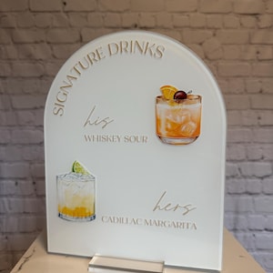 Może przedstawiać: Biały akrylowy znak z łukowatym wierzchem, z napisem "Signature Drinks" i ilustracjami koktajli. Zawiera "his" Whiskey Sour i "hers" Cadillac Margarita. Przezroczysta akrylowa podstawa.