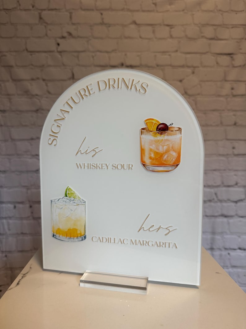 Może przedstawiać: Biały akrylowy znak z łukowatym wierzchem, z napisem "Signature Drinks" i ilustracjami koktajli. Zawiera "his" Whiskey Sour i "hers" Cadillac Margarita. Przezroczysta akrylowa podstawa.
