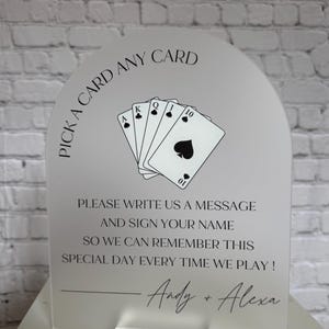 May include: An arched frosted acrylic sign featuring a black graphic of five playing cards and the text: PICK A CARD ANY CARD, PLEASE WRITE US A MESSAGE AND SIGN YOUR NAME SO WE CAN REMEMBER THIS SPECIAL DAY EVERY TIME WE PLAY! Andy + Alexa.