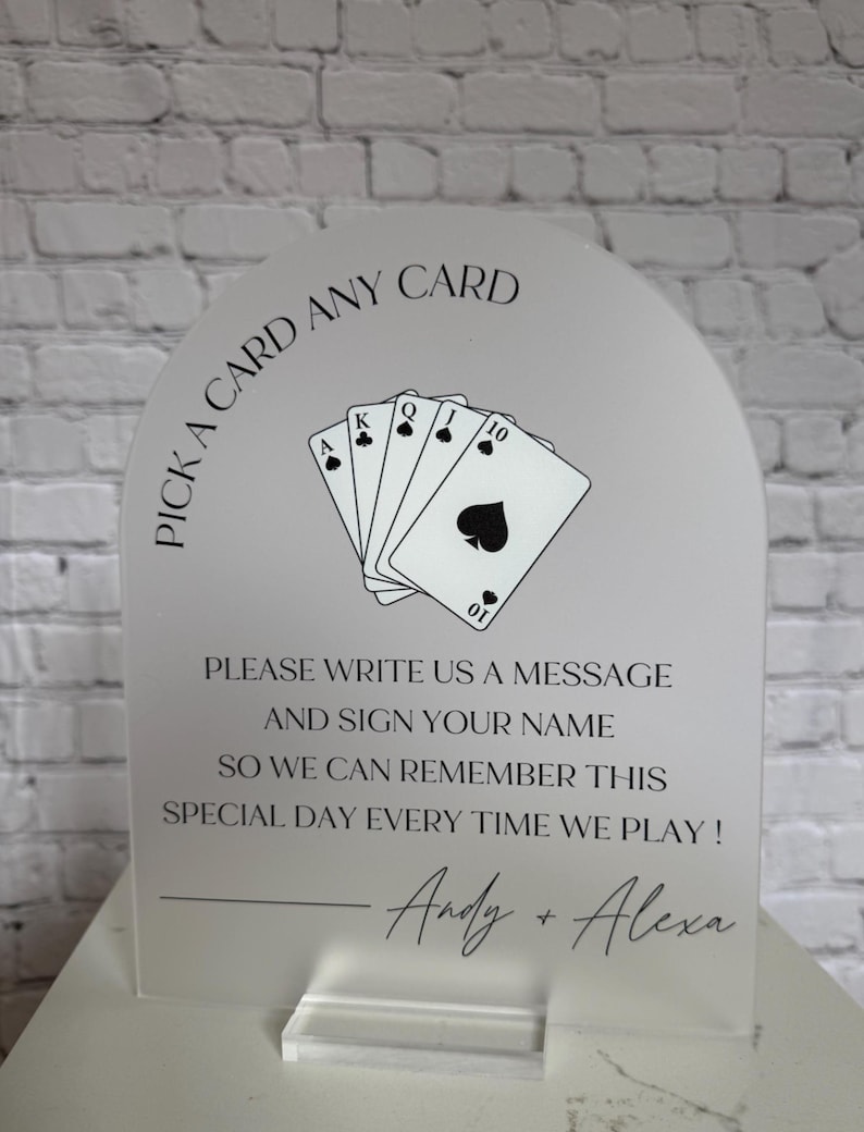 May include: An arched frosted acrylic sign featuring a black graphic of five playing cards and the text: PICK A CARD ANY CARD, PLEASE WRITE US A MESSAGE AND SIGN YOUR NAME SO WE CAN REMEMBER THIS SPECIAL DAY EVERY TIME WE PLAY! Andy + Alexa.