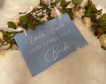 May include: A light blue sign with white text that reads "Uncle Hunter, HERE COMES YOUR Bride." The sign has white ribbon handles and is surrounded by a floral garland with white and cream-colored flowers.