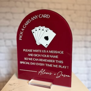 May include: An arched, deep red acrylic sign featuring a white graphic of a royal flush of spades playing cards. The text reads: PICK A CARD ANY CARD, PLEASE WRITE US A MESSAGE AND SIGN YOUR NAME SO WE CAN REMEMBER THIS SPECIAL DAY EVERY TIME WE PLAY! Alexis + Jason.
