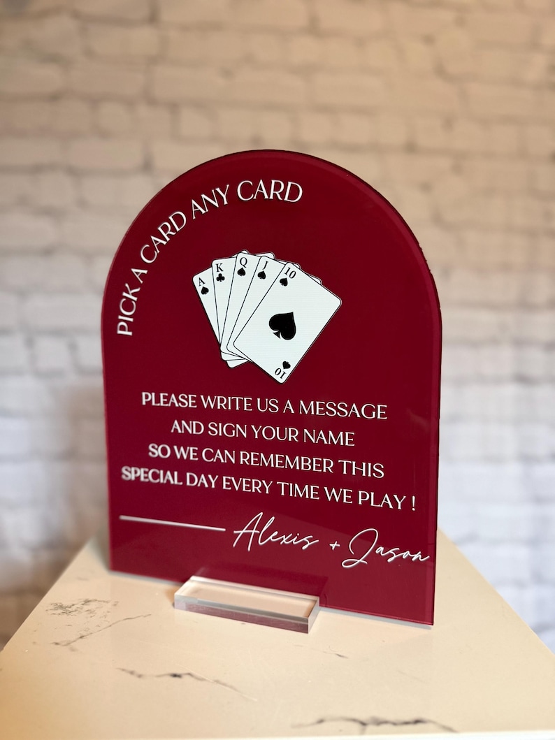 May include: An arched, deep red acrylic sign featuring a white graphic of a royal flush of spades playing cards. The text reads: PICK A CARD ANY CARD, PLEASE WRITE US A MESSAGE AND SIGN YOUR NAME SO WE CAN REMEMBER THIS SPECIAL DAY EVERY TIME WE PLAY! Alexis + Jason.