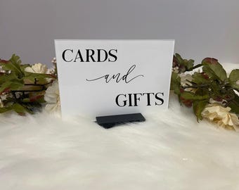 May include: White acrylic sign with black text that reads "CARDS and GIFTS". The sign is on a white fluffy surface.