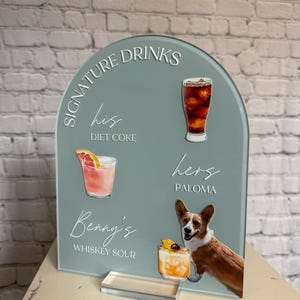 May include: A teal acrylic drinks sign with the words "Signature Drinks" and drink illustrations. The sign includes the text "his Diet Coke", "hers Paloma", and "Benny's Whiskey Sour". A corgi dog is featured on the sign.