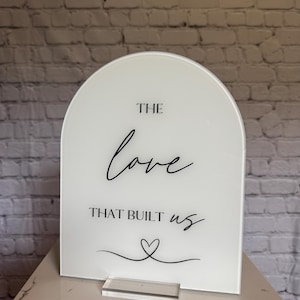 May include: A white acrylic sign with an arched top. The sign features the words "THE love THAT BUILT us" in black script, along with a heart symbol. It stands on a clear acrylic base.