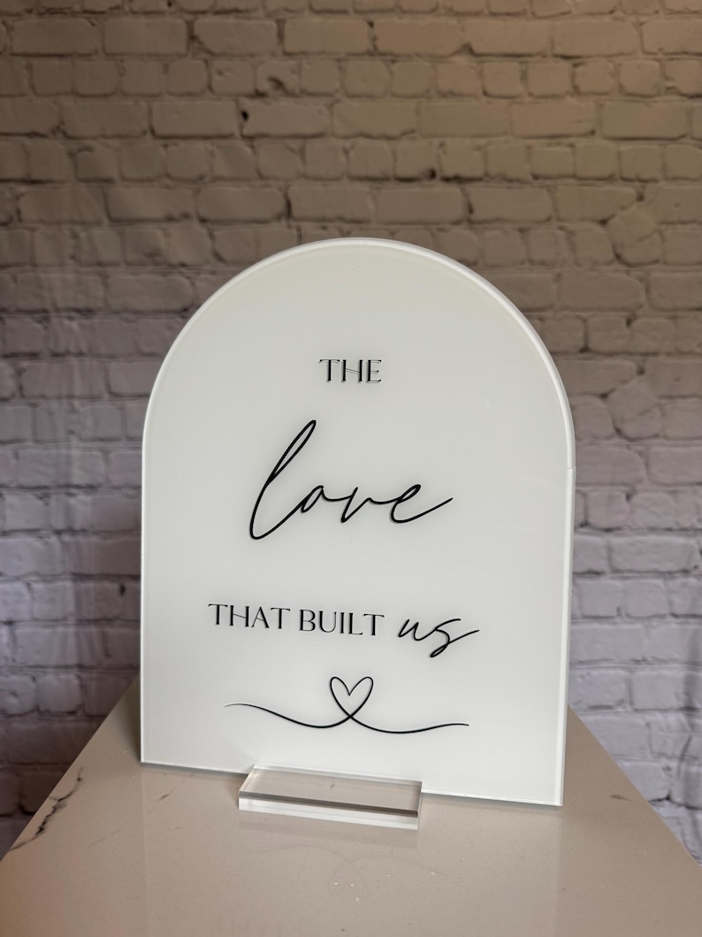 May include: A white acrylic sign with an arched top. The sign features the words "THE love THAT BUILT us" in black script, along with a heart symbol. It stands on a clear acrylic base.