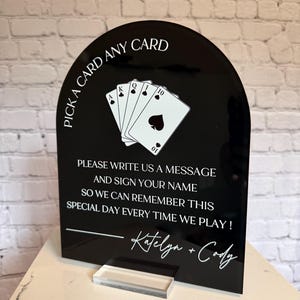 May include: A black arched acrylic sign with white text and a graphic of four playing cards. The text reads: PICK A CARD ANY CARD, PLEASE WRITE US A MESSAGE AND SIGN YOUR NAME SO WE CAN REMEMBER THIS SPECIAL DAY EVERY TIME WE PLAY! Katelyn + Cody.