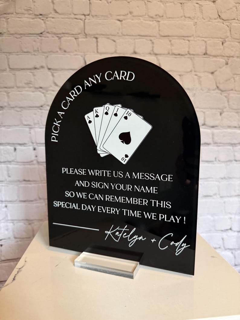 May include: A black arched acrylic sign with white text and a graphic of four playing cards. The text reads: PICK A CARD ANY CARD, PLEASE WRITE US A MESSAGE AND SIGN YOUR NAME SO WE CAN REMEMBER THIS SPECIAL DAY EVERY TIME WE PLAY! Katelyn + Cody.