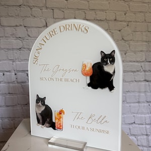 May include: White acrylic sign with gold text reading "Signature Drinks". It features illustrations of cocktails and black and white cats. The sign lists "The Grayson" and "The Bella" cocktails.