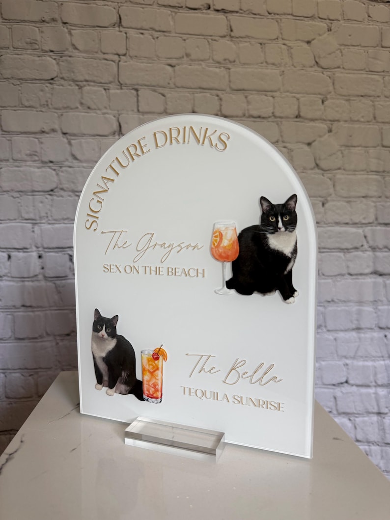 May include: White acrylic sign with gold text reading "Signature Drinks". It features illustrations of cocktails and black and white cats. The sign lists "The Grayson" and "The Bella" cocktails.