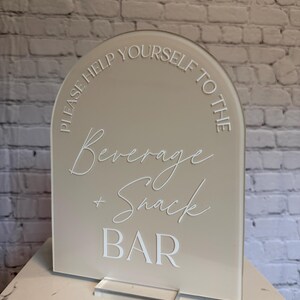 May include: An arched beige acrylic sign with white lettering that reads: PLEASE HELP YOURSELF TO THE Beverage + Snack BAR. The sign is mounted on a clear rectangular acrylic base, featuring a combination of sans-serif and script fonts.