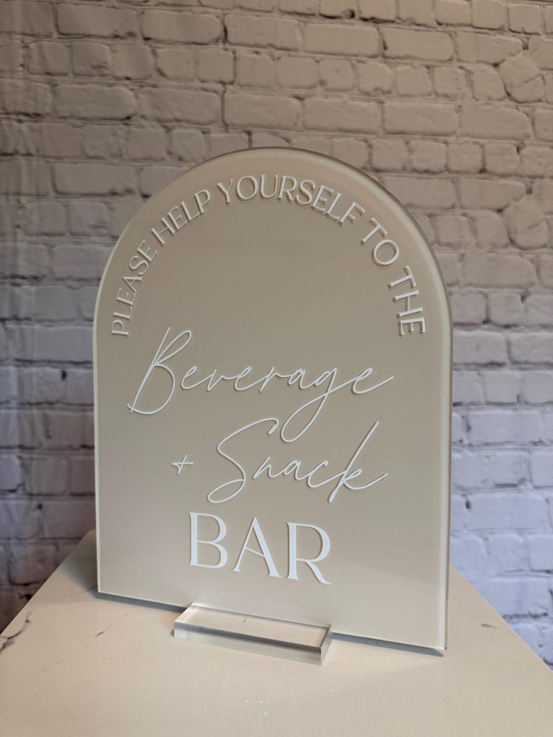 May include: An arched beige acrylic sign with white lettering that reads: PLEASE HELP YOURSELF TO THE Beverage + Snack BAR. The sign is mounted on a clear rectangular acrylic base, featuring a combination of sans-serif and script fonts.