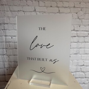 May include: A frosted acrylic sign with the words "THE love THAT BUILT us" in black script. A small heart design is below the text. The sign is displayed on a clear acrylic stand, set against a white brick wall.