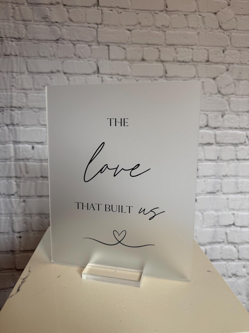 May include: A frosted acrylic sign with the words "THE love THAT BUILT us" in black script. A small heart design is below the text. The sign is displayed on a clear acrylic stand, set against a white brick wall.