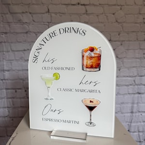 May include: White acrylic sign with an arched top, featuring the words "Signature Drinks" and illustrations of cocktails: Old Fashioned, Classic Margarita, and Espresso Martini. Includes a clear acrylic stand.