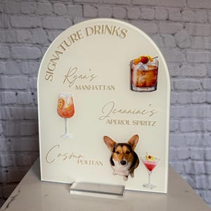 May include: A cream acrylic drinks sign with the words "Signature Drinks" and cocktail names. The sign features illustrations of a Manhattan, Aperol Spritz, and Cosmopolitan, plus a corgi dog.