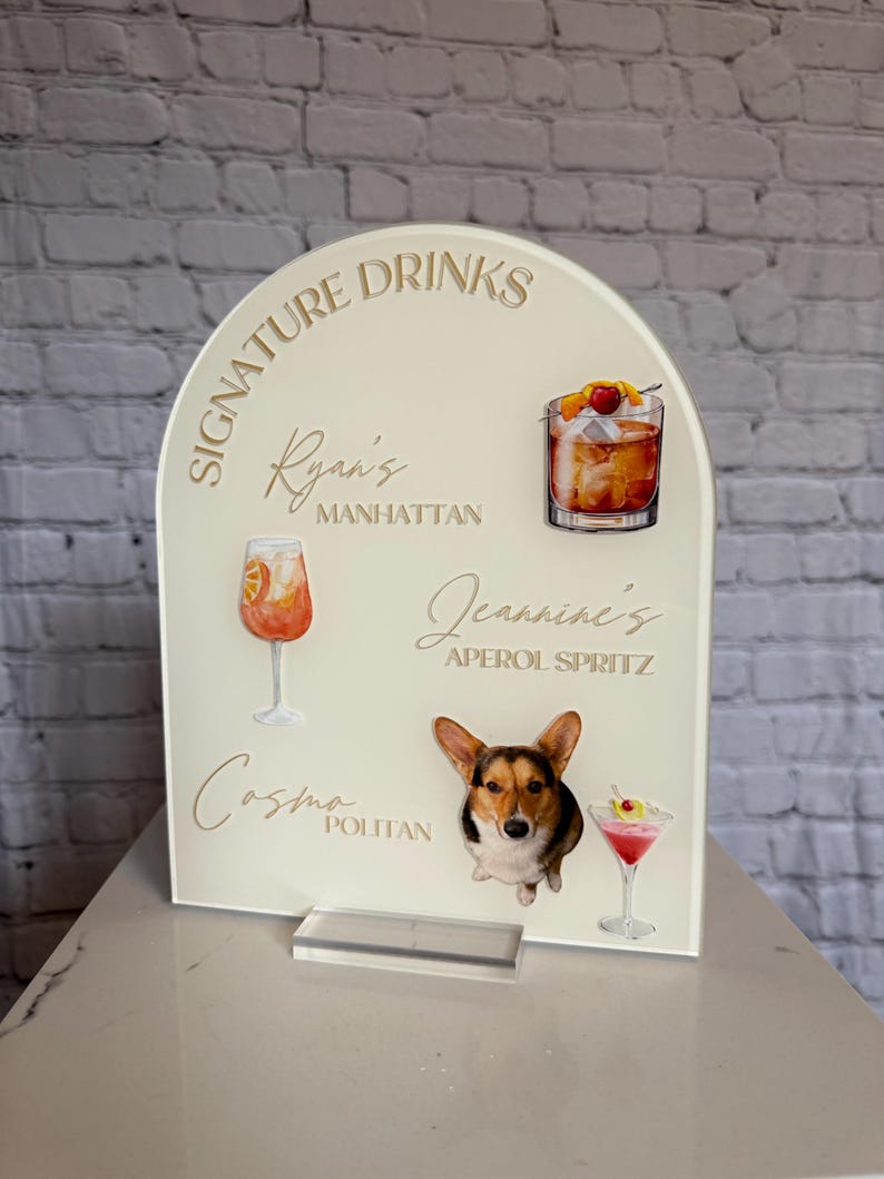 May include: A cream acrylic drinks sign with the words "Signature Drinks" and cocktail names. The sign features illustrations of a Manhattan, Aperol Spritz, and Cosmopolitan, plus a corgi dog.