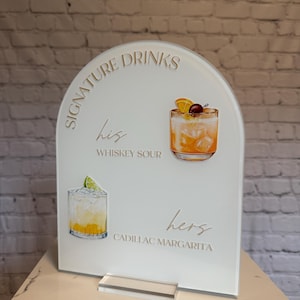 Może przedstawiać: Biały akrylowy znak z napisem "Signature Drinks" i ilustracjami koktajli. Znak zawiera tekst "his Whiskey Sour" i "hers Cadillac Margarita". Znak jest łukowaty u g&oacute;ry i ma przezroczystą akrylową podstawę.