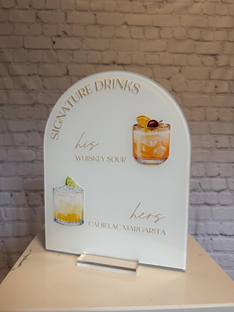 Może przedstawiać: Biały akrylowy znak z napisem "Signature Drinks" i ilustracjami koktajli. Znak zawiera tekst "his Whiskey Sour" i "hers Cadillac Margarita". Znak jest łukowaty u g&oacute;ry i ma przezroczystą akrylową podstawę.