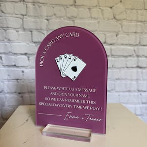 May include: An arched, deep magenta acrylic sign featuring a white graphic of a hand of playing cards and the text: PICK A CARD ANY CARD, PLEASE WRITE US A MESSAGE AND SIGN YOUR NAME SO WE CAN REMEMBER THIS SPECIAL DAY EVERY TIME WE PLAY! Emma + Tanner.