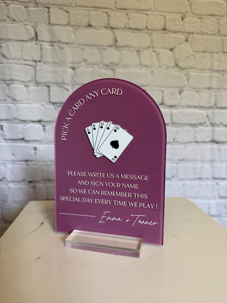 May include: An arched, deep magenta acrylic sign featuring a white graphic of a hand of playing cards and the text: PICK A CARD ANY CARD, PLEASE WRITE US A MESSAGE AND SIGN YOUR NAME SO WE CAN REMEMBER THIS SPECIAL DAY EVERY TIME WE PLAY! Emma + Tanner.