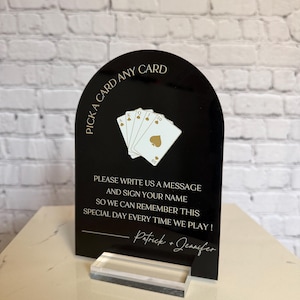 May include: An arched black acrylic sign with gold text and graphics. The top reads PICK A CARD ANY CARD above an illustration of four playing cards. Below is the text PLEASE WRITE US A MESSAGE AND SIGN YOUR NAME SO WE CAN REMEMBER THIS SPECIAL DAY EVERY TIME WE PLAY! followed by Patrick + Jennifer. The sign sits on a clear acrylic base.