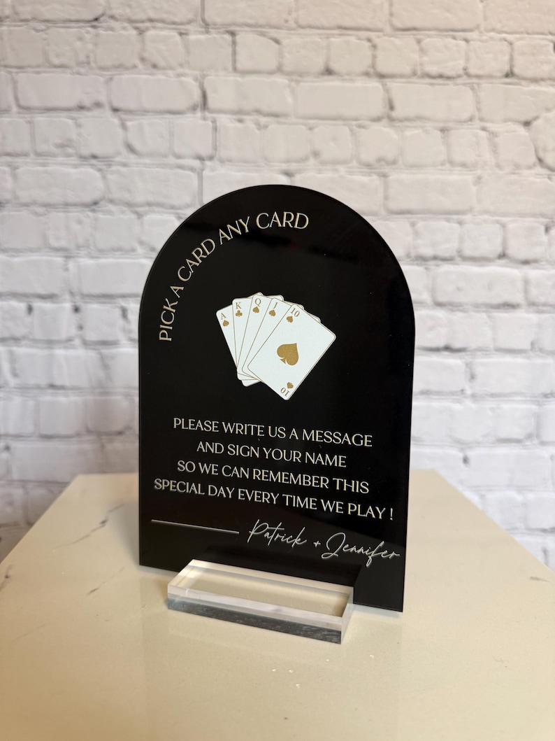 May include: An arched black acrylic sign with gold text and graphics. The top reads PICK A CARD ANY CARD above an illustration of four playing cards. Below is the text PLEASE WRITE US A MESSAGE AND SIGN YOUR NAME SO WE CAN REMEMBER THIS SPECIAL DAY EVERY TIME WE PLAY! followed by Patrick + Jennifer. The sign sits on a clear acrylic base.