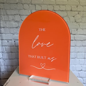May include: An orange acrylic sign with an arched top. White script reads "THE love THAT BUILT us" with a heart symbol. The sign is displayed on a clear acrylic base, suitable for weddings or special events.