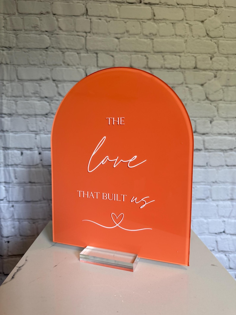May include: An orange acrylic sign with an arched top. White script reads "THE love THAT BUILT us" with a heart symbol. The sign is displayed on a clear acrylic base, suitable for weddings or special events.