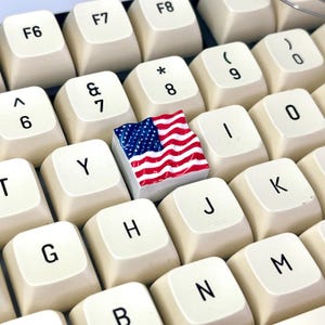 May include: A standard keyboard with a small American flag keycap replacing the letter 'I' key. The flag is red, white, and blue.