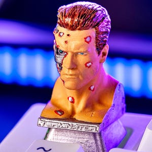 May include: A bust of the Terminator cyborg, with detailed facial features and exposed mechanical components. The bust is tan with red accents and brown hair. The base has the word "TERMINATOR" on it. The bust is placed on a white keyboard.