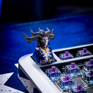 May include: A silver mechanical keyboard with a custom keycap featuring a detailed 3D printed bust of a female figure with horns and red eyes. The keyboard has clear keycaps with purple switches.
