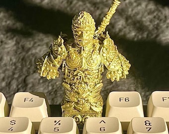 May include: A gold-colored figurine of a monkey warrior wearing armor and holding a sword. The figurine is sitting on a keyboard.