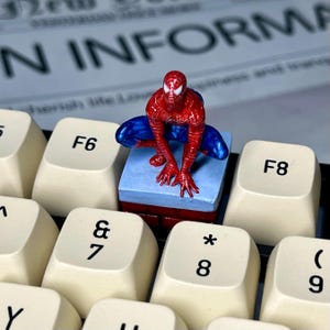 May include: A red and blue Spider-Man figurine is perched on a small brick and blue platform, positioned on a white keyboard with black keys. The keys feature the letters F6, F8, and numbers 7, 8, and 9.