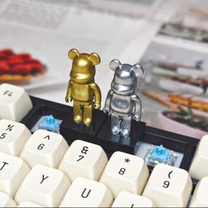 May include: Two small bear-shaped figurines, one gold and one silver, stand on a black base between the keys of a white keyboard. The keyboard keys are labeled with black letters and symbols.