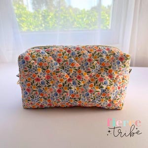 May include: A quilted cosmetic bag with a floral pattern in shades of pink, blue, yellow, and orange. The bag has a rectangular shape with a zip closure and the text "fierce tribe".