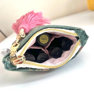 Op de afbeelding: Een kleine, groene stoffen etui met een gouden rits en een roze kwast. De etui is open en onthult verschillende kleine, donkere flesjes. Eén flesje is gelabeld met "Lime". De etui is ontworpen om etherische olie flesjes in te bewaren.