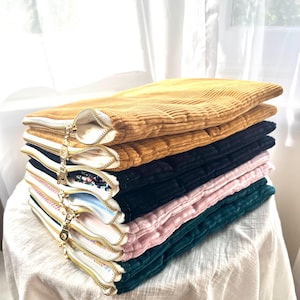 Könnte beinhalten: Ein Stapel Cord-Reißverschlusstaschen in verschiedenen Farben, darunter Gold, Schwarz, Rosa und Türkis. Jede Tasche hat einen goldenen Reißverschluss und ein weißes Futter. Die Taschen sind übereinander gestapelt.