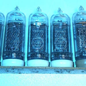 Puede incluir: Una fila de siete tubos nixie de vidrio transparente montados sobre una placa de circuito rectangular. Cada tubo contiene una serie de números de alambre metálico apilados y una rejilla, con bases circulares blancas.