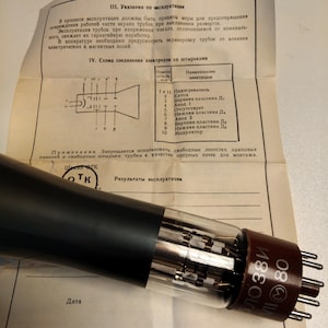 Puede incluir: Una válvula de vacío vintage con una base marrón y múltiples pines, parcialmente encerrada en un tubo de vidrio transparente. El tubo está colocado sobre un documento con texto y diagramas cirílicos. El texto incluye las palabras "Resultados de la operación."