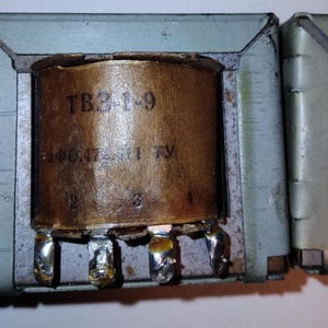 Puede incluir: Un componente transformador eléctrico con un núcleo de bobina envuelto en papel marrón con el texto TB3-1-9 y marcas alfanuméricas adicionales impresas en la superficie. Tiene cuatro terminales de soldadura metálicos y un marco gris.