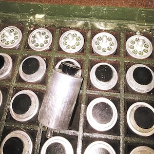 Puede incluir: Una colección de componentes eléctricos antiguos, que incluye piezas cilíndricas plateadas y piezas circulares con centros negros, cuidadosamente dispuestas en una caja de madera verde. Algunos componentes tienen pequeñas bombillas transparentes.