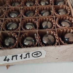 May include: A cardboard box with a grid of individual compartments, each holding a small, reflective, spherical object. The objects appear to be made of a dark, metallic material. The box has a label with the text "4П1Л⊕".