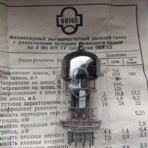 Puede incluir: Un tubo de vacío transparente con el texto "6H14П" impreso. El tubo tiene una parte superior gris oscuro y múltiples pines en la parte inferior. Está sobre un papel impreso con texto cirílico.