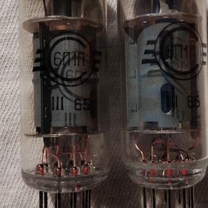 Può includere: Due valvole termoioniche vintage con bulbi in vetro trasparente e basi metalliche. Ogni valvola ha una forma cilindrica con componenti interni visibili. Le valvole presentano testo e marcature nere, tra cui "6П1П" e "65".