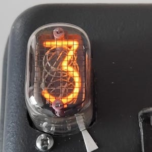 May include: A close-up of a vintage Nixie tube clock displaying the number "3" in a bright orange glow. The tube is encased in glass and has a metal frame.