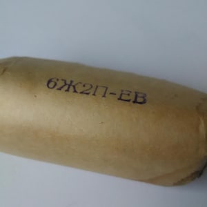 Peut inclure: Objet cylindrique enveloppé dans du papier beige. Le texte "6Ж2П-EB" est estampillé à l'encre bleu foncé sur le papier. L'objet est légèrement incurvé et semble être scellé.