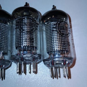 Può includere: Primo piano di tre valvole termoioniche in vetro con componenti interni argentati e più pin alla base. I tubi hanno una parte superiore arrotondata, su sfondo bianco.